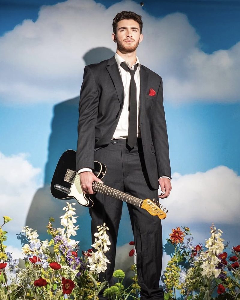 Joe Ricci in a suit holding an electric guitar among wildflowers